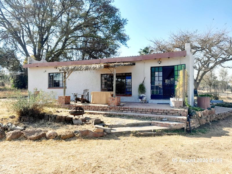 Rear view of a farm cottage featuring a brick patio, thatched pergola, and established garden surroundings.
