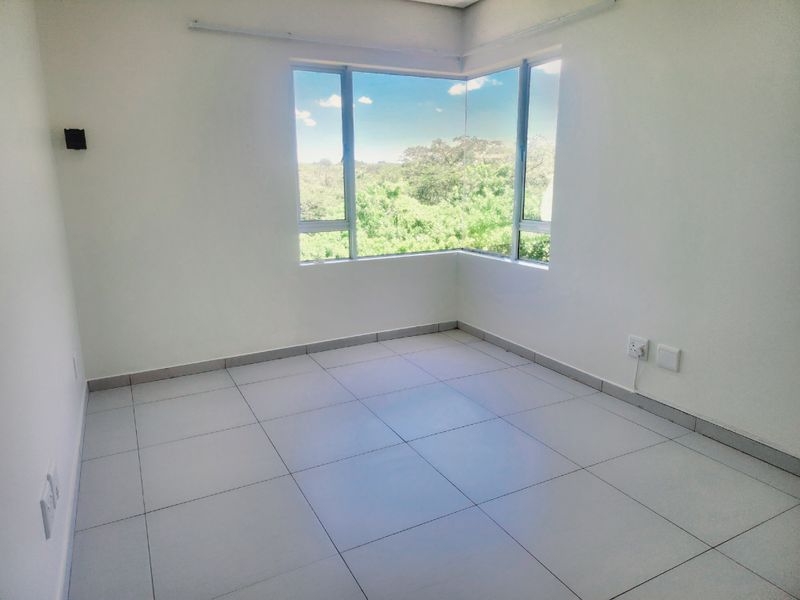 A bright, empty bedroom featuring large corner windows with natural views, neutral tiled flooring, and clean white walls.