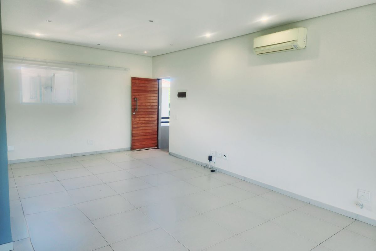 A bright, empty living area featuring neutral tiled flooring, recessed downlighting, and a wall-mounted air conditioning unit, providing a clean and versatile space.