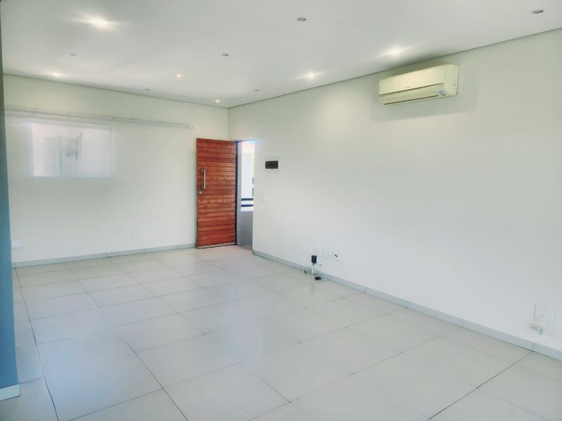 A bright, empty living area featuring neutral tiled flooring, recessed downlighting, and a wall-mounted air conditioning unit, providing a clean and versatile space.