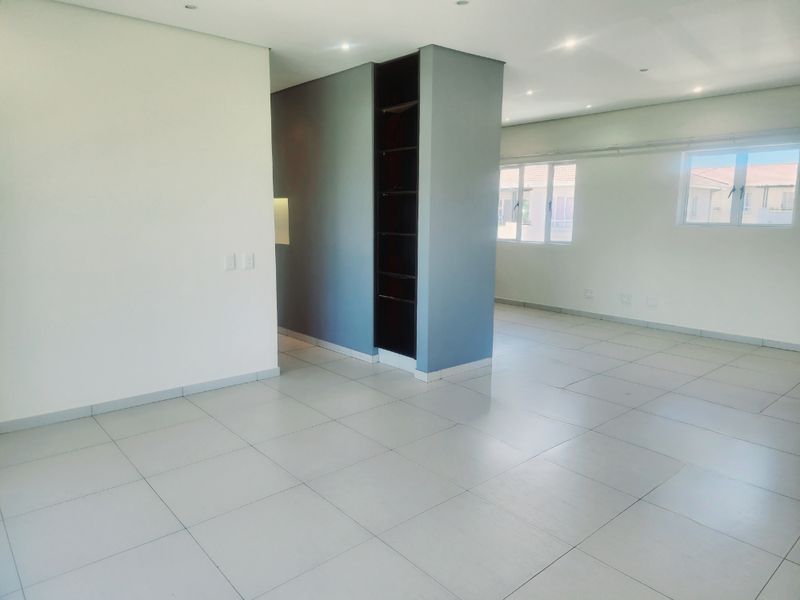 A bright, open-plan living area featuring neutral tiled flooring, recessed downlighting, and a modern built-in shelving unit, offering a versatile and spacious layout.