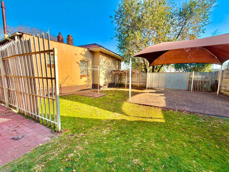 Rear garden area featuring a paved patio with a shade sail, lawn, and perimeter security fencing.