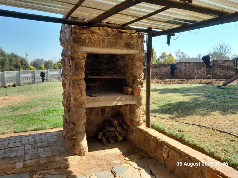 Outdoor patio area featuring a rustic stone built-in braai, paved flooring, and a covered roof structure overlooking the garden.