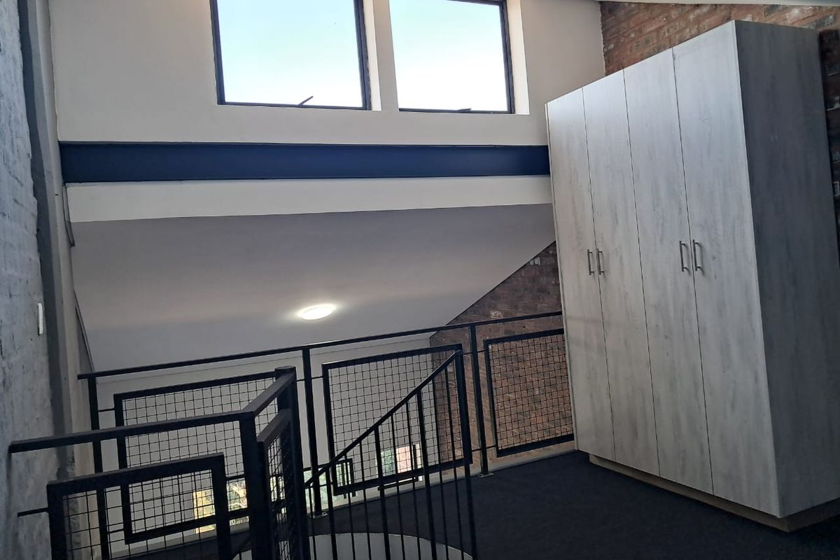 A loft-style bedroom area featuring exposed brickwork, a modern metal staircase railing, built-in wardrobe storage, and high clerestory windows providing natural light.