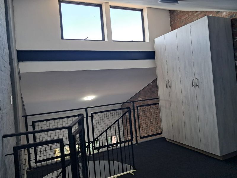 A loft-style bedroom area featuring exposed brickwork, a modern metal staircase railing, built-in wardrobe storage, and high clerestory windows providing natural light.