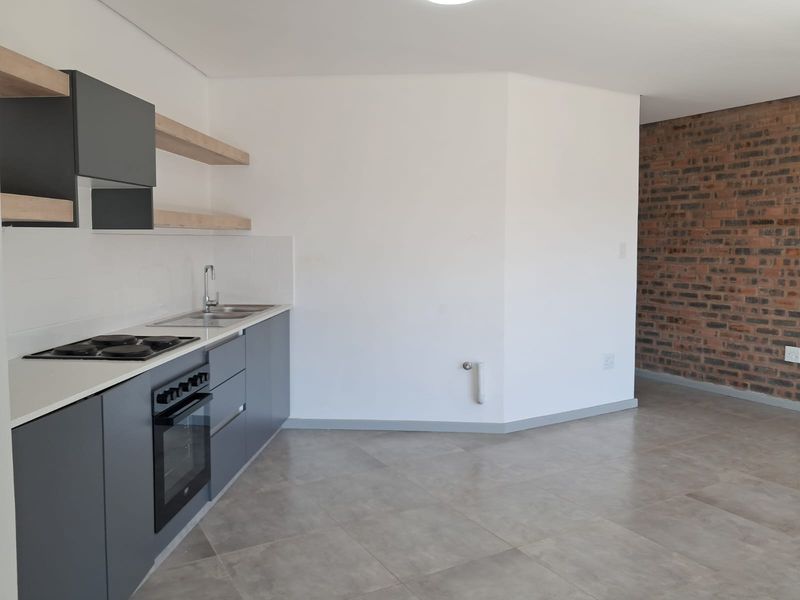 Modern open-plan kitchen and living area featuring sleek grey cabinetry, built-in appliances, tiled floors, and a striking exposed brick feature wall.