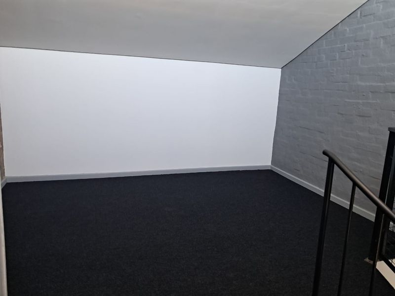An empty loft-style bedroom featuring a sloped ceiling, dark carpeting, and an exposed grey brick wall with a metal staircase railing.