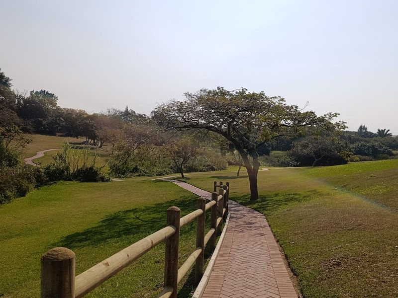 A well-maintained paved walkway winds through a landscaped garden area, featuring timber post and rail fencing and established trees.