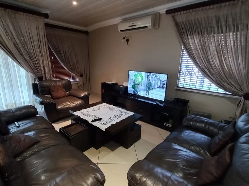 A comfortable living room featuring tiled flooring, a wall-mounted air conditioning unit, and large windows fitted with security bars and curtains.
