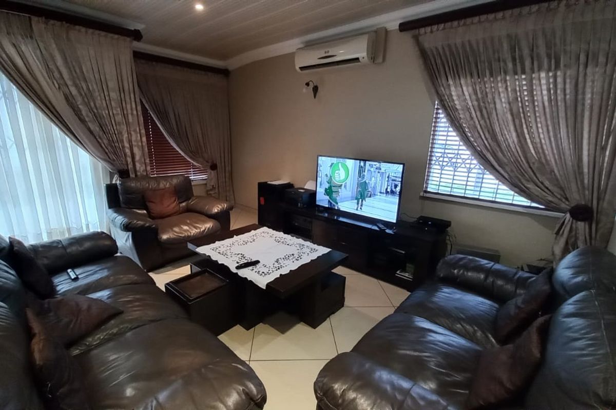 A comfortable living room featuring tiled flooring, a wall-mounted air conditioning unit, and large windows fitted with security bars and curtains.