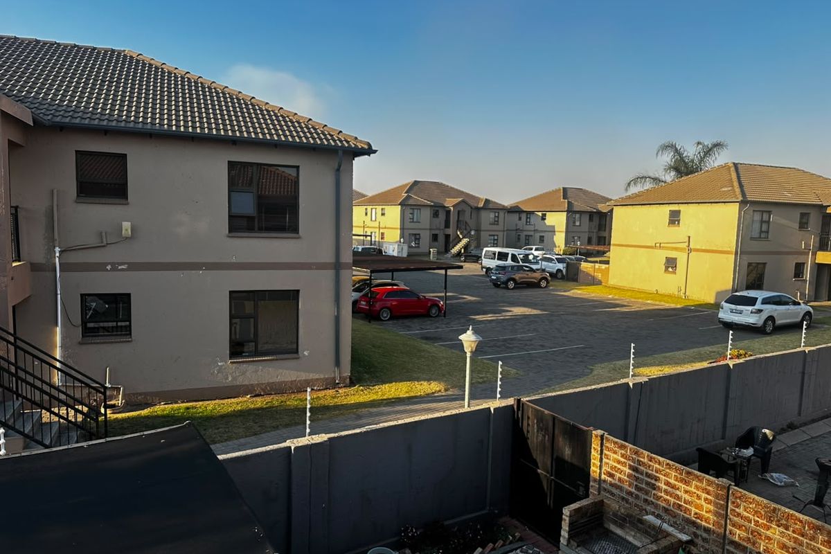 Rear view of the townhouse complex featuring a paved parking area, carport, brick boundary wall with electric fencing, and a built-in brick braai.