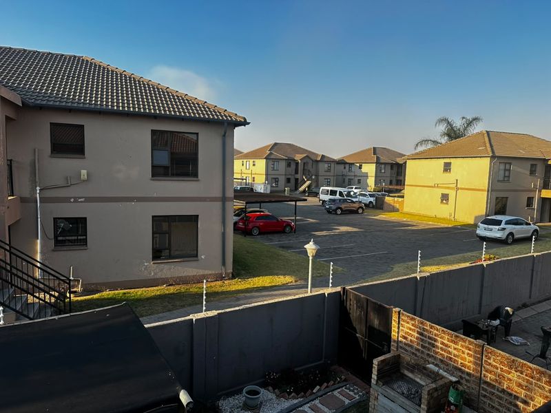 Rear view of the townhouse complex featuring a paved parking area, carport, brick boundary wall with electric fencing, and a built-in brick braai.