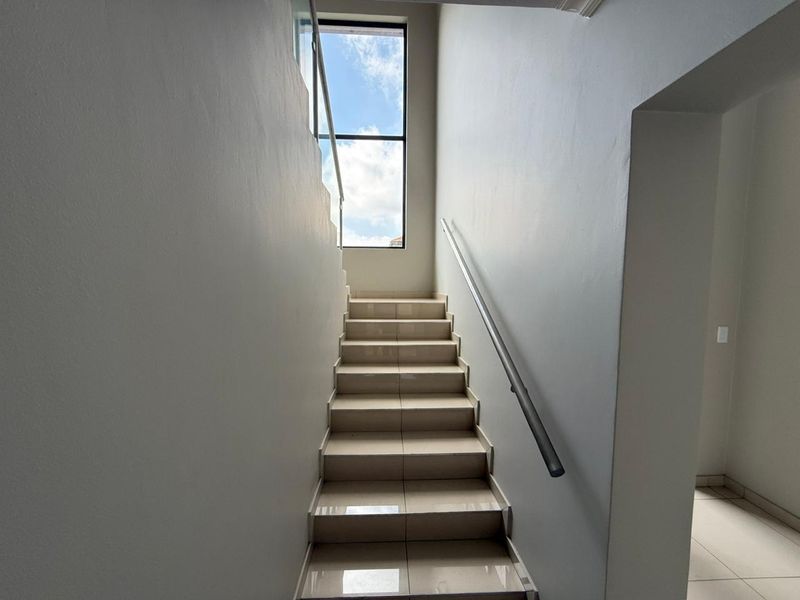 A bright, modern staircase featuring tiled steps, a sleek metal handrail, and a glass balustrade, illuminated by a large window.