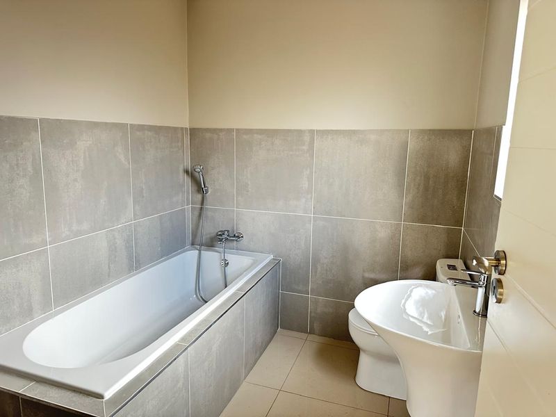 A clean, modern bathroom featuring a built-in bathtub with handheld shower mixer, a pedestal basin, and a toilet with neutral grey wall tiling.