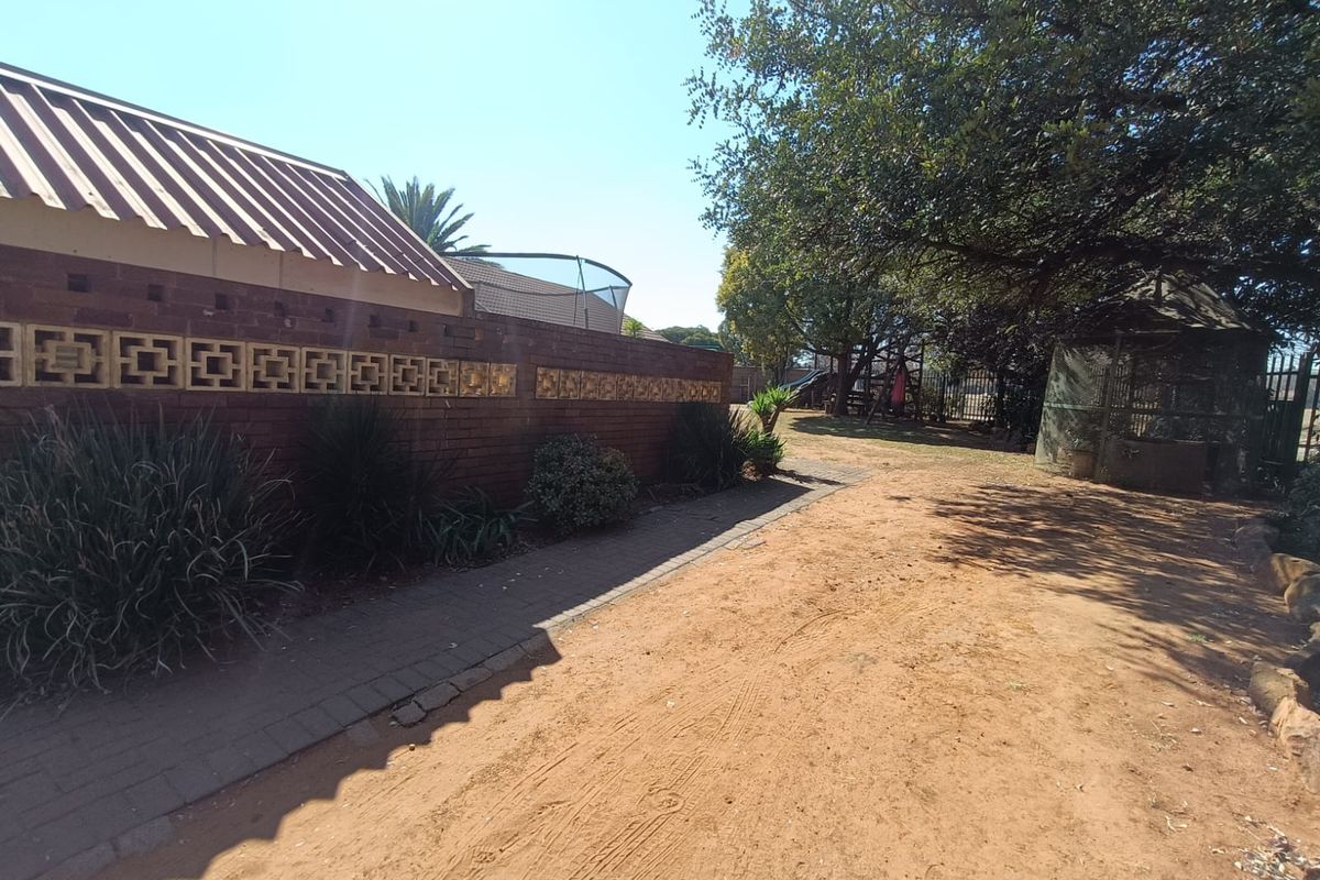 A rear exterior view featuring a brick boundary wall with decorative breeze blocks, a paved walkway, and a large dirt driveway area.