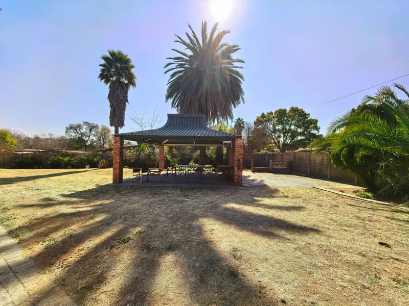A spacious outdoor entertainment area featuring a covered patio with brick pillars and a metal roof, set within a large garden.