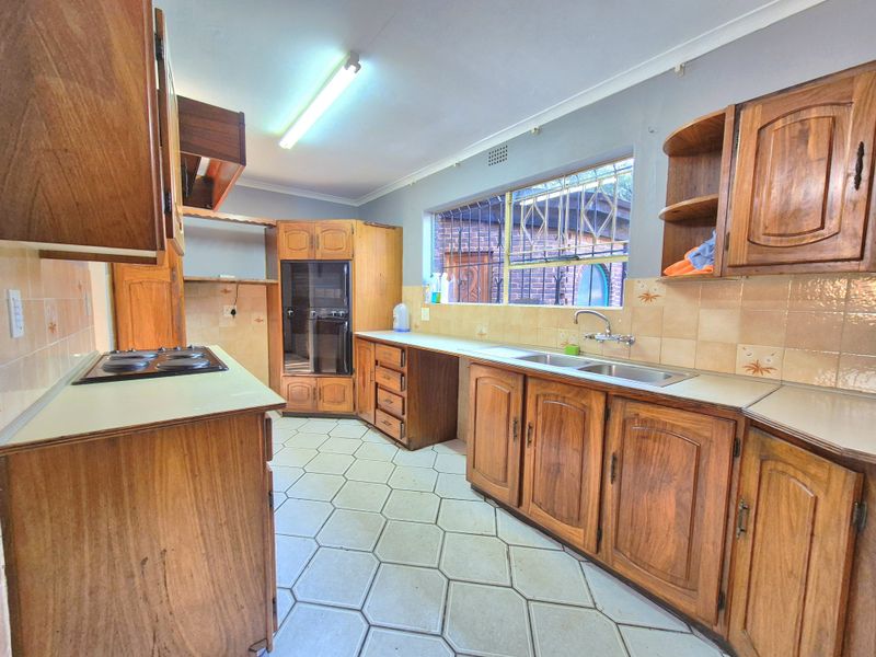 A functional kitchen featuring wooden cabinetry, built-in oven, electric hob, and double sink with tiled backsplash and flooring.