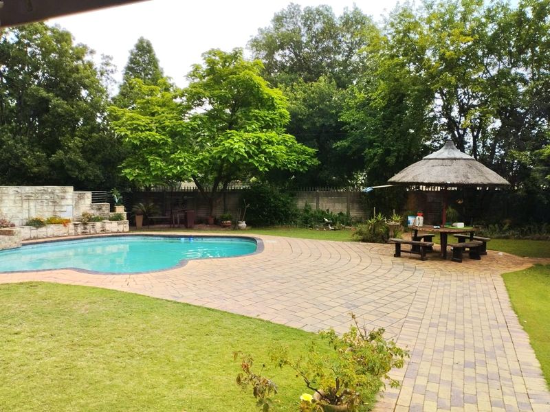 Spacious outdoor pool area featuring a paved deck, stone retaining wall, and a traditional thatched lapa, set within a mature garden.