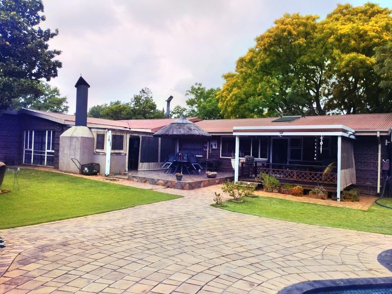 Rear view of the property featuring a brick facade, covered patio area, outdoor fireplace, paved courtyard, and a swimming pool set within a garden.
