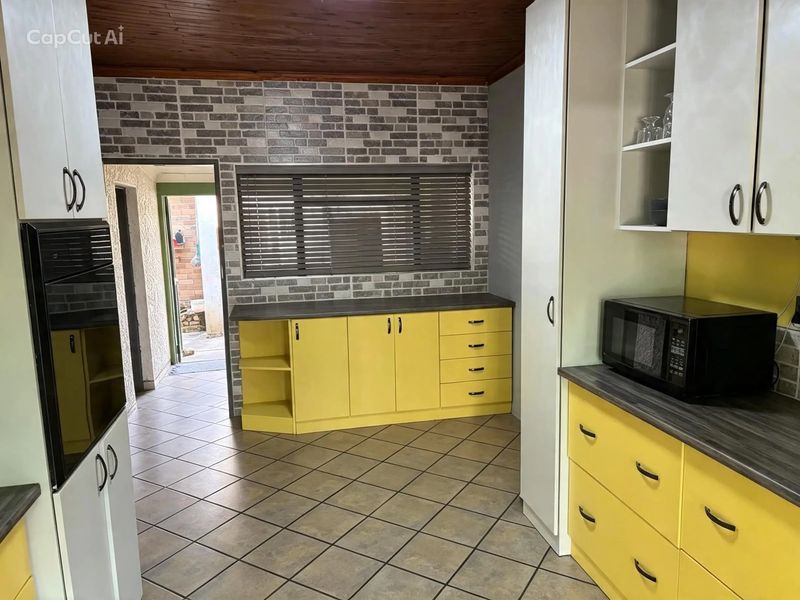 A kitchen featuring yellow cabinetry, dark laminate countertops, tiled flooring, and a timber ceiling, with a brick-effect feature wall and access to an exterior area.