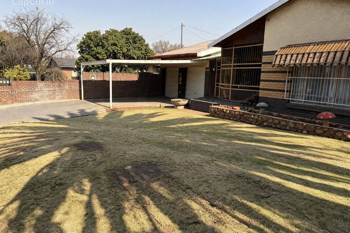 Rear exterior view featuring a lawn, paved driveway, covered patio area, and brick boundary wall with security features.