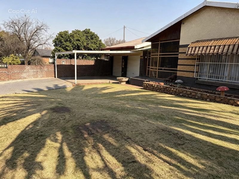 Rear exterior view featuring a lawn, paved driveway, covered patio area, and brick boundary wall with security features.
