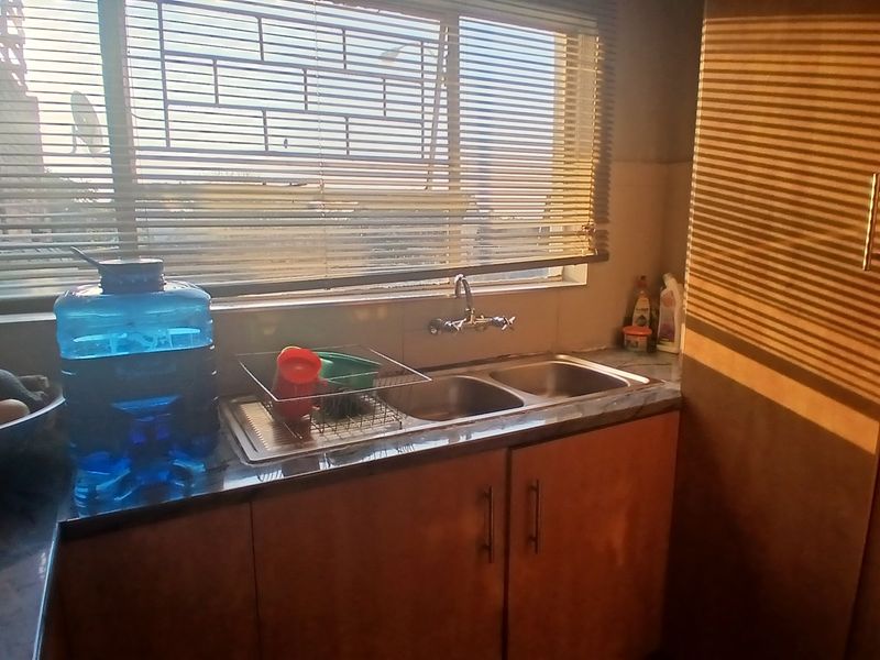 A functional kitchen area featuring a double bowl stainless steel sink, laminate cabinetry, and a countertop, illuminated by natural light through venetian blinds.