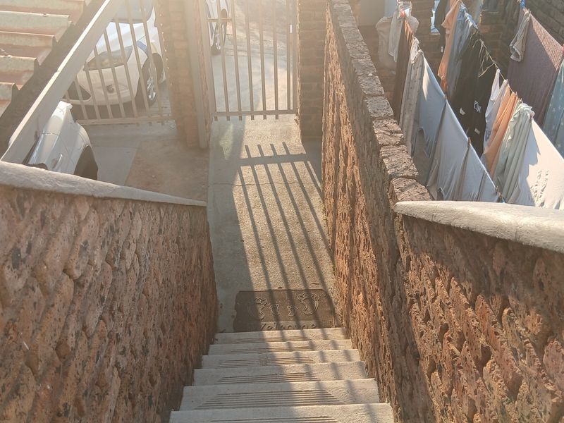 An exterior view of a concrete staircase leading to a gated entrance, flanked by brick boundary walls and showing signs of weathering.