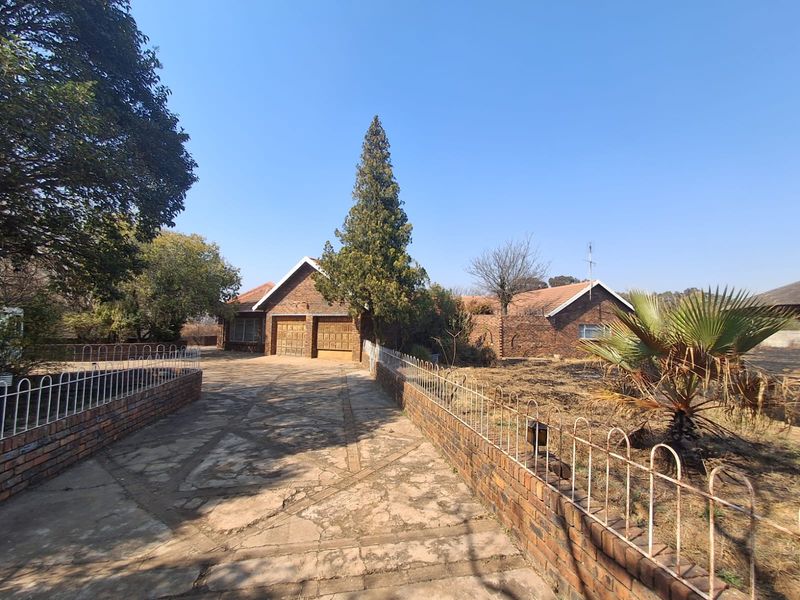 Front exterior view of a brick house featuring a double garage, paved driveway, and perimeter walling with metal fencing in a mature garden setting.
