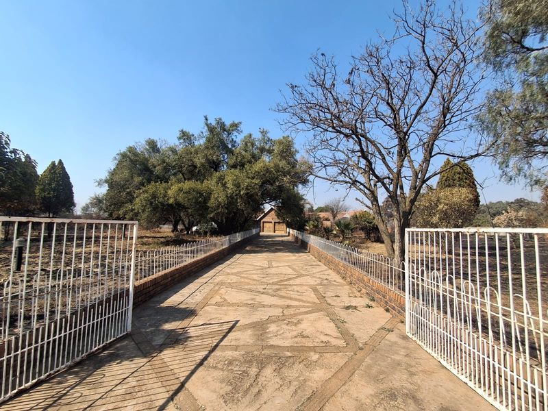 A long paved driveway leading to a garage, framed by metal gates and mature trees on a large property.