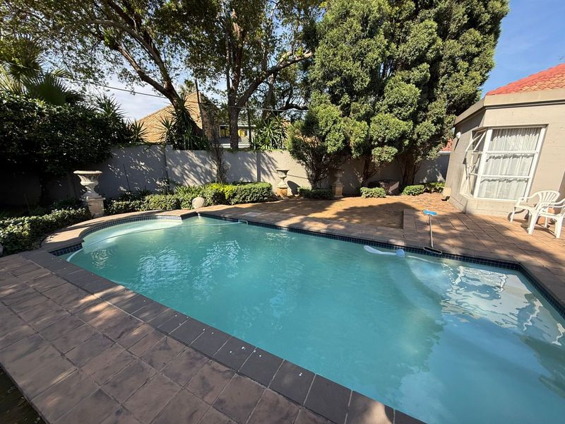 A well-maintained outdoor pool area featuring a tiled surround, paved patio space, and established garden boundary walls.