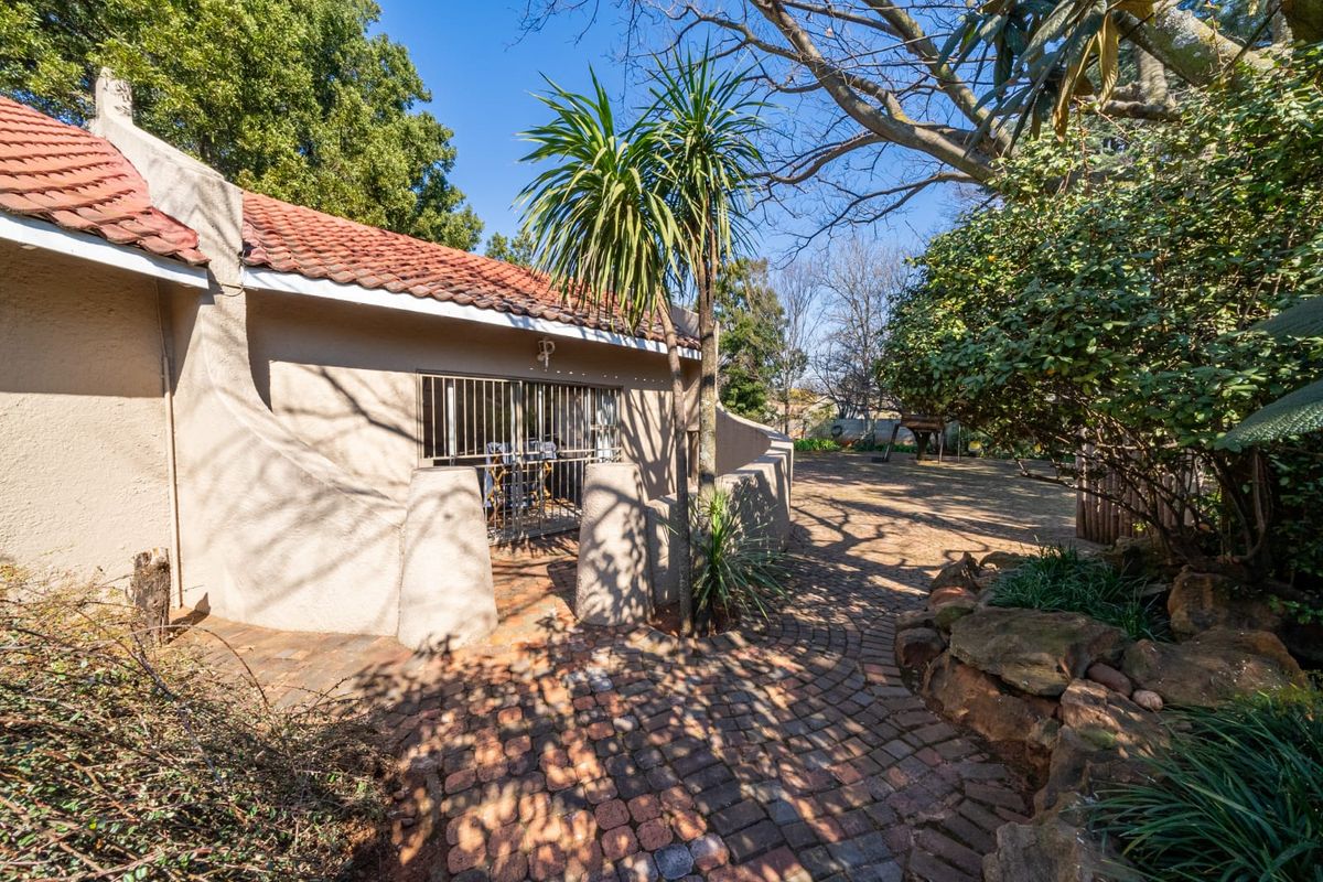 A rear exterior view of the house featuring a terracotta tiled roof, stucco walls, and a brick-paved patio area surrounded by established garden landscaping.