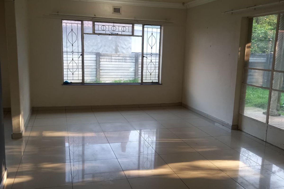An empty, sunlit living room featuring tiled flooring, security bars on the windows, and direct access to the garden through a sliding glass door.