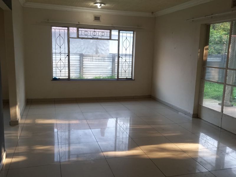 An empty, sunlit living room featuring tiled flooring, security bars on the windows, and direct access to the garden through a sliding glass door.