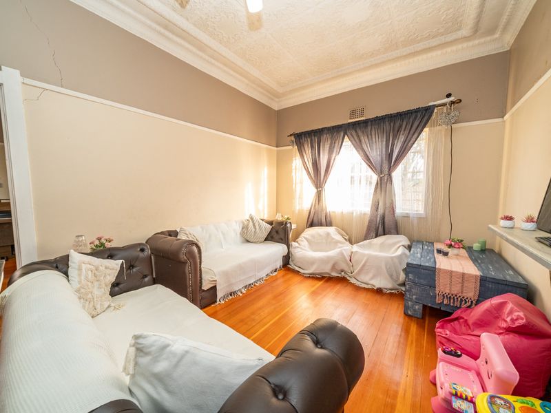 A living room featuring original hardwood flooring, ornate pressed metal ceilings, and a fireplace mantel, showing signs of wear and wall cracking.