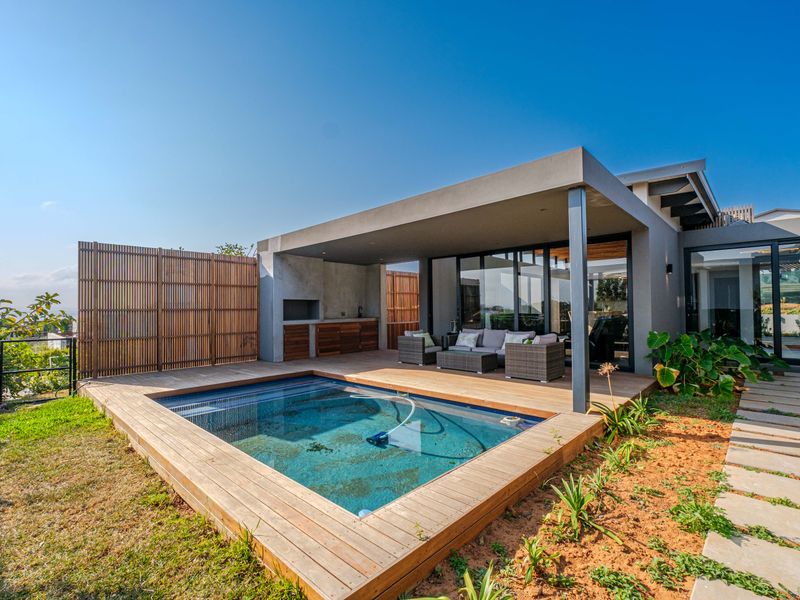 Modern outdoor entertainment area featuring a swimming pool, timber decking, covered patio with built-in braai, and seamless indoor-outdoor flow through large sliding doors.