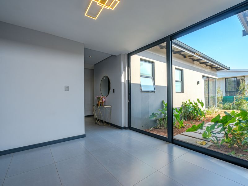 A bright, modern hallway featuring large format floor tiles, contemporary lighting, and expansive sliding glass doors that provide seamless access to the outdoor garden area.