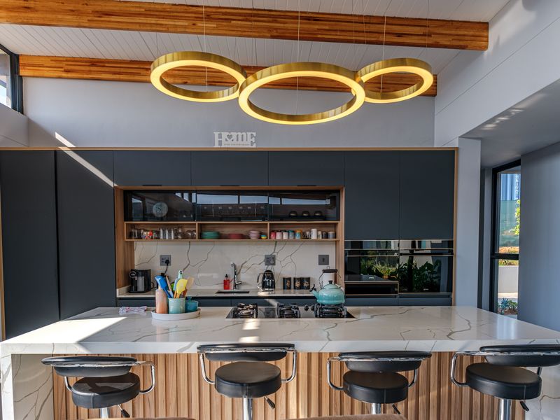 Modern kitchen featuring dark cabinetry, a large quartz island with integrated hob, marble-look splashback, and striking circular pendant lighting under timber-beamed ceilings.