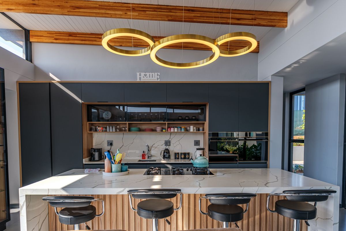 Modern kitchen featuring dark cabinetry, a large quartz island with integrated hob, marble-look splashback, and striking circular pendant lighting under timber-beamed ceilings.