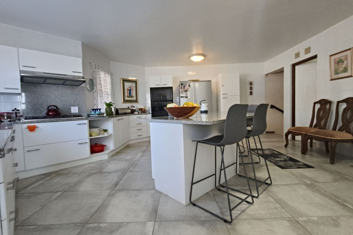 A bright, open-plan kitchen featuring white cabinetry, granite countertops, a central island with breakfast bar seating, and neutral tiled flooring.