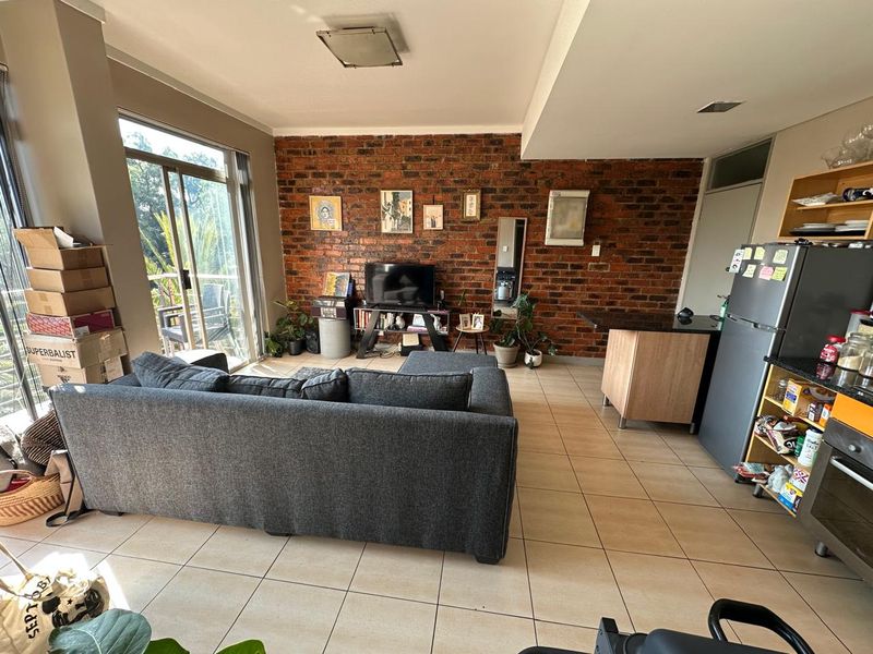Open-plan living area featuring an exposed brick wall, tiled flooring, and direct access to a balcony, seamlessly integrated with a functional kitchen space.