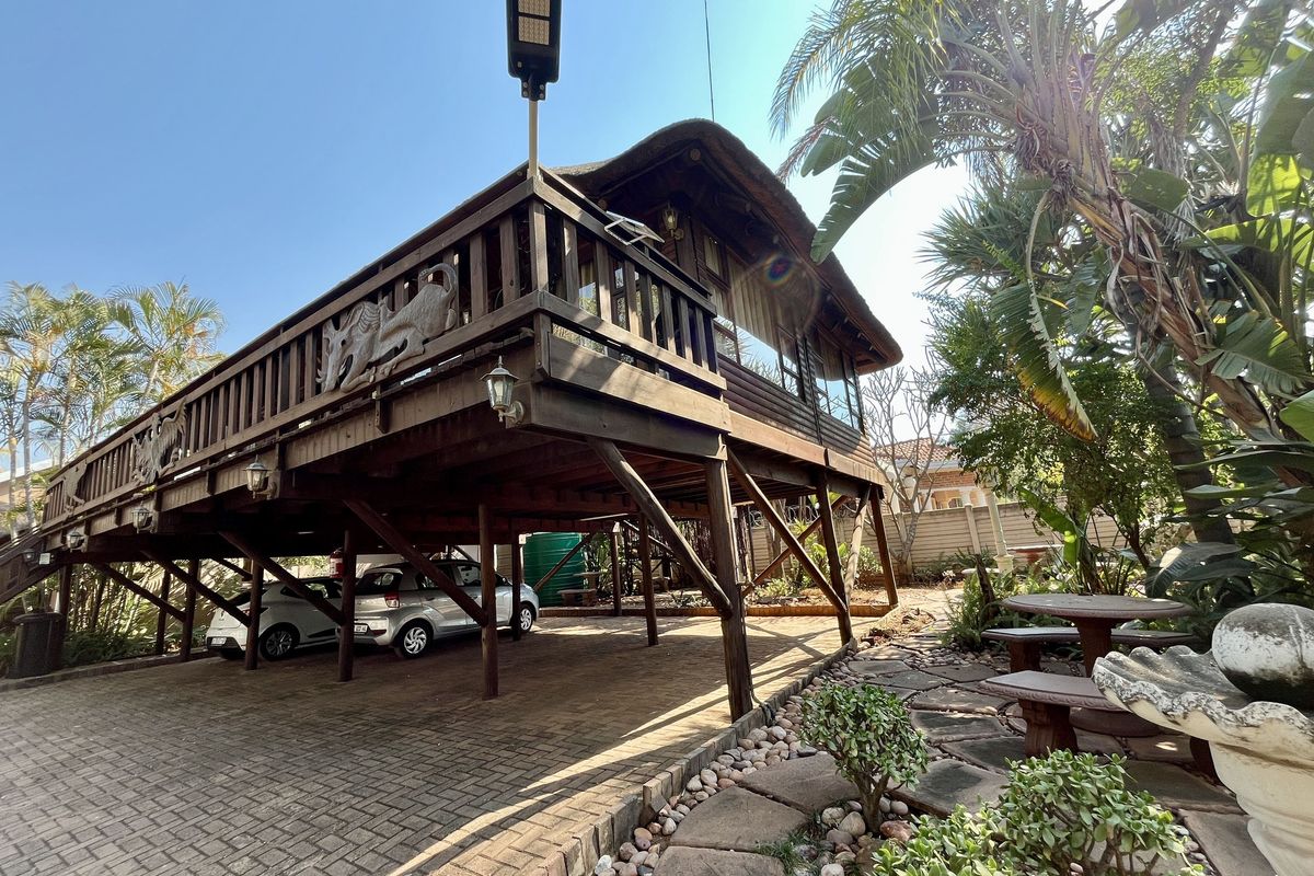 Elevated timber deck with a thatch roof, featuring a paved driveway underneath and a landscaped garden area.