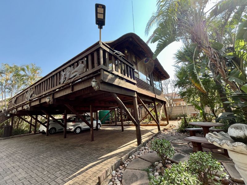 Elevated timber deck with a thatch roof, featuring a paved driveway underneath and a landscaped garden area.