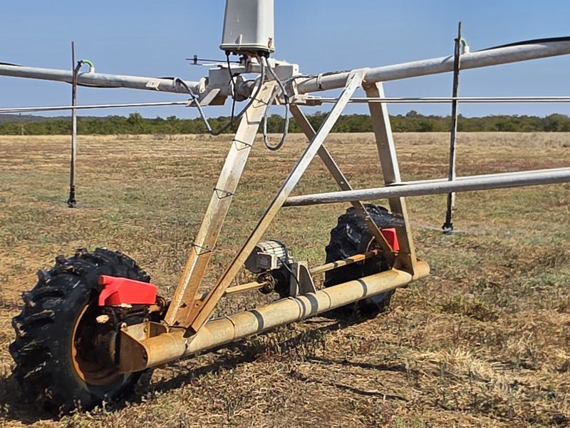 A center pivot irrigation system operating in a large agricultural field, featuring metal support structures and sprinkler heads for crop watering.