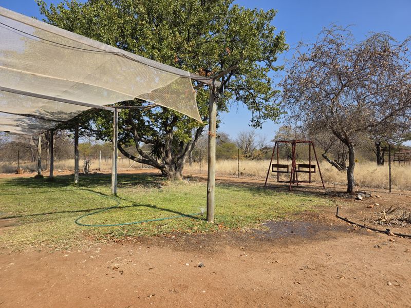 A rural garden area featuring a shade netting structure supported by wooden posts, set within a natural farm landscape with perimeter fencing.