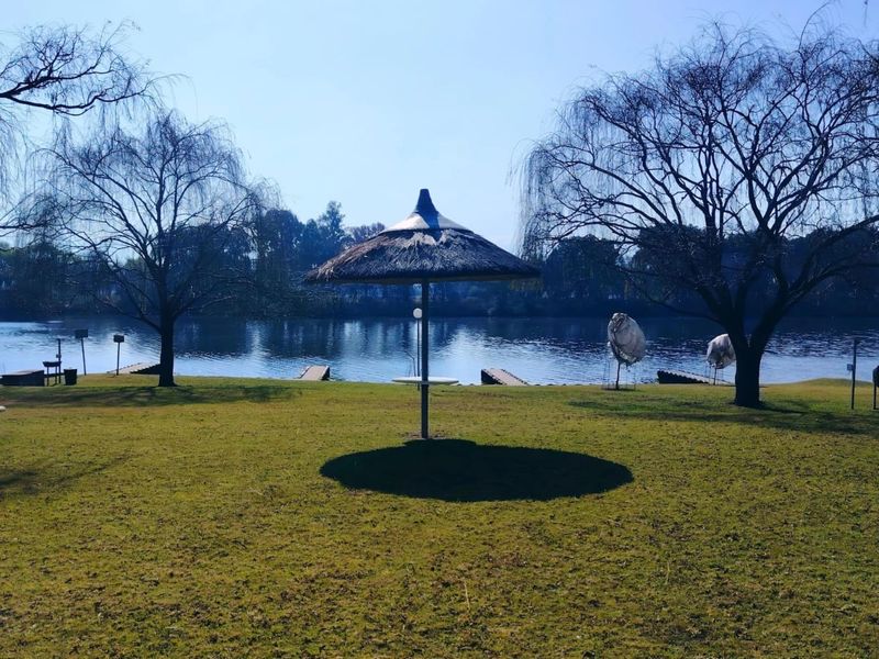 A scenic view of a tranquil lakefront area featuring a manicured lawn, mature trees, and a thatched umbrella structure.