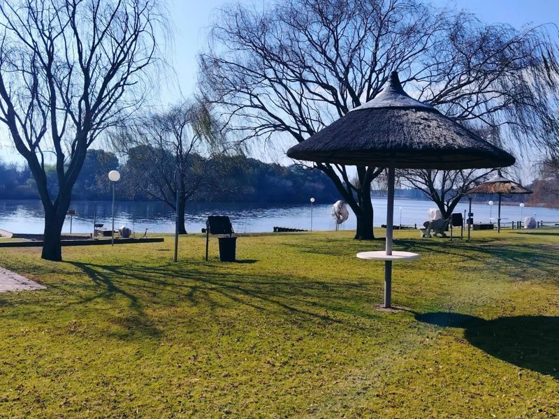 A scenic riverfront garden area featuring established trees, manicured lawns, and fixed thatched umbrellas, offering direct access to the water.