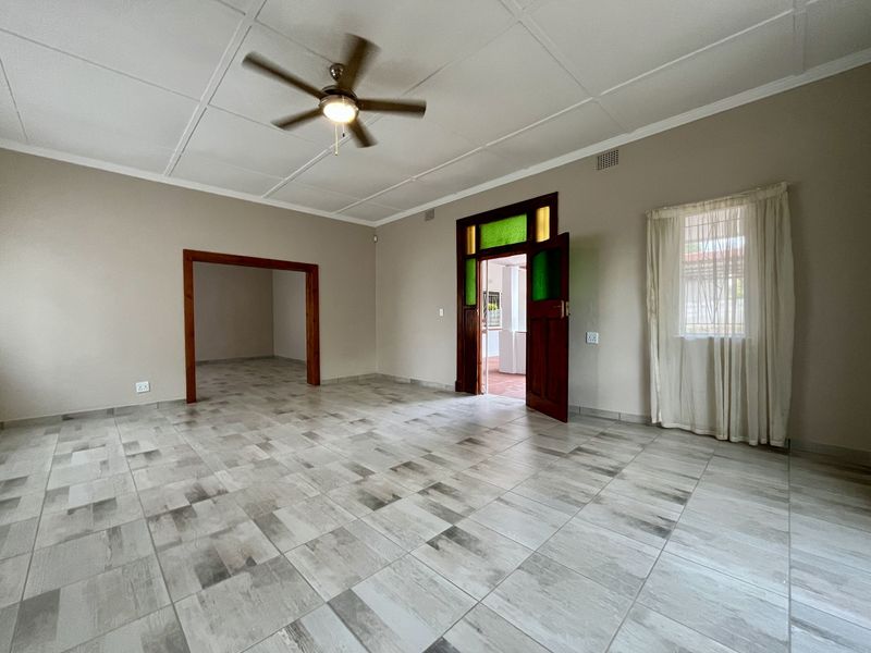 A spacious, empty living room featuring neutral tiled flooring, a ceiling fan, and a characterful timber door with stained glass accents.