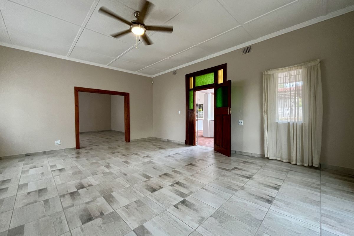 A spacious, empty living room featuring neutral tiled flooring, a ceiling fan, and a characterful timber door with stained glass accents.