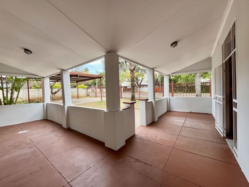 A spacious covered patio area featuring painted concrete flooring, structural pillars, and direct access to the front garden and driveway.